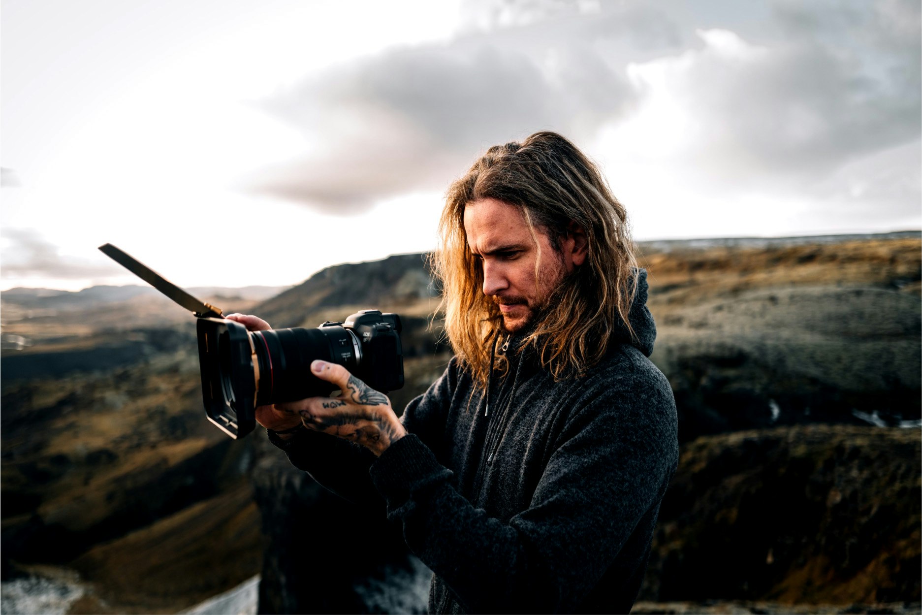 A content creator with long hair focuses on his camera, capturing the scenic beauty of an outdoor landscape, perfect for stock footage