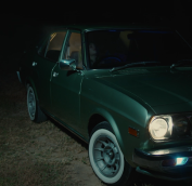 A dark green vintage car, parked with one light on.