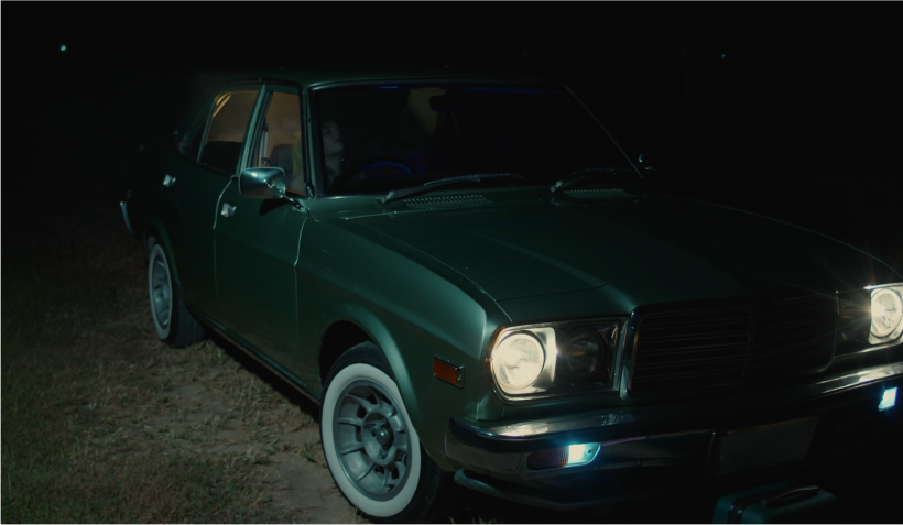A dark green vintage car, parked with one light on.